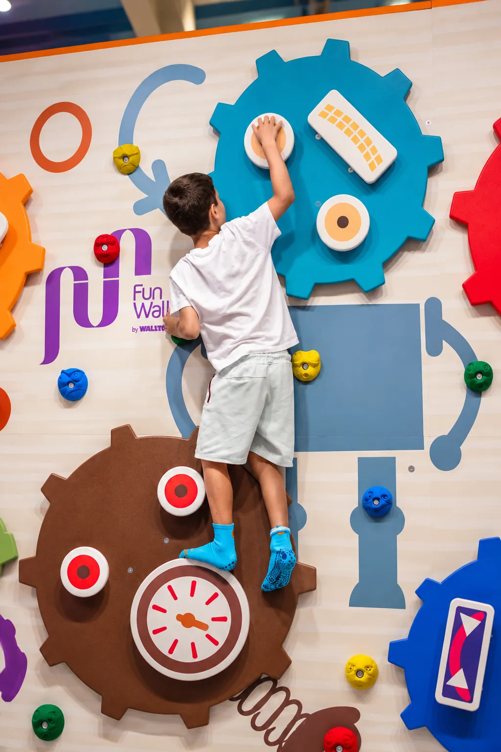 Băiat cățărându-se pe peretele de escaladă Fun Wall cu roți dințate colorate din zona KLANkids Adventure