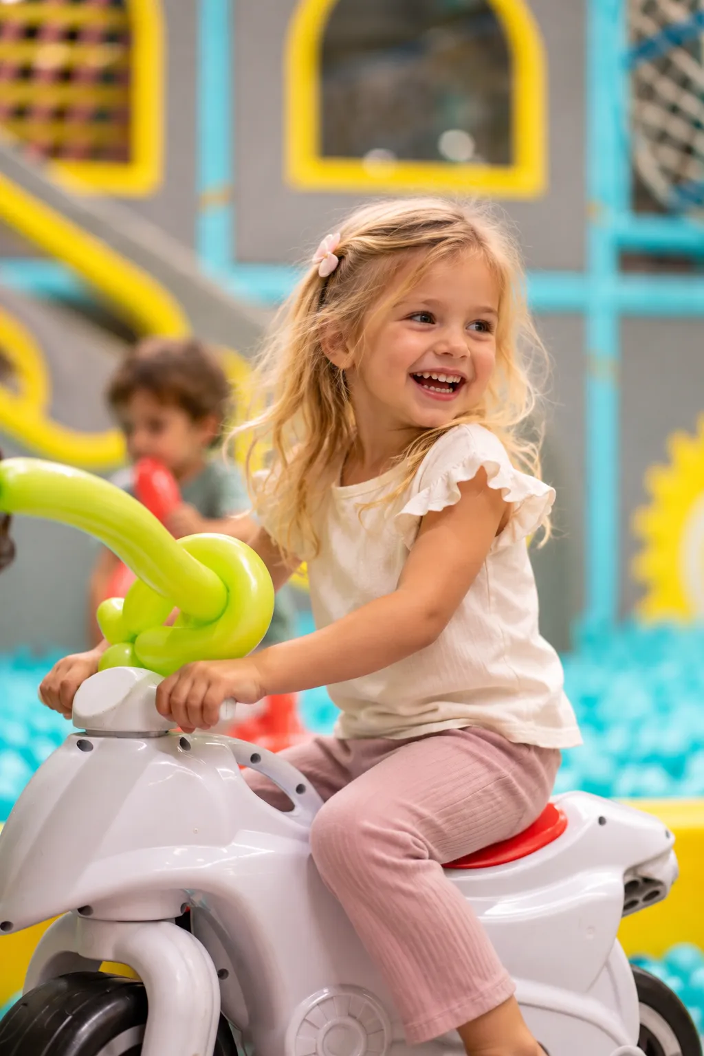 Fetiță zâmbitoare pe o motocicletă de jucărie în zona KLANkids Carnaval, cu piscina cu bile în fundal