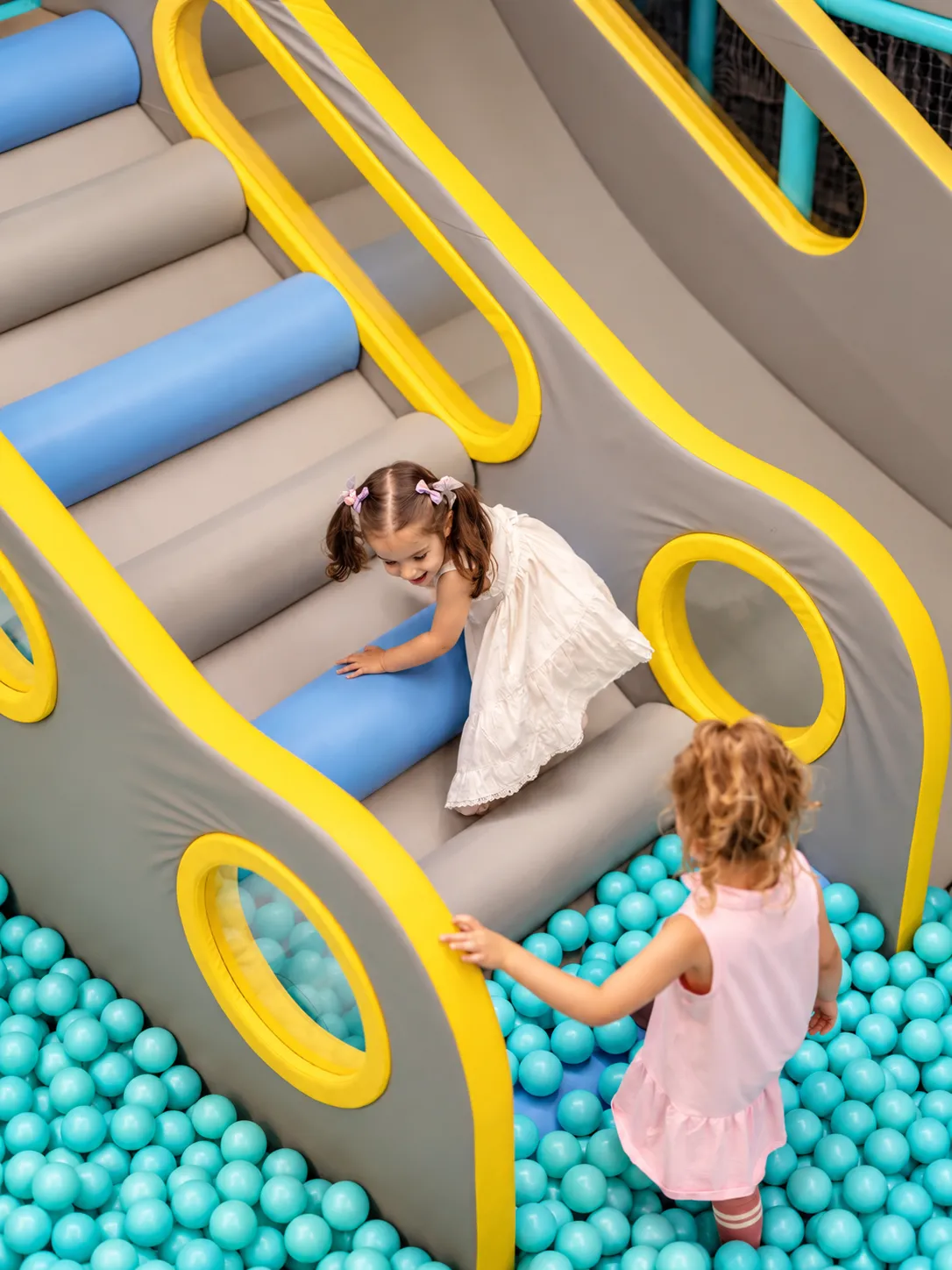 Două fetițe jucându-se împreună pe structura gri-galbenă cu role albastre, deasupra piscinei cu bile turcoaz din KLANkids Carnaval