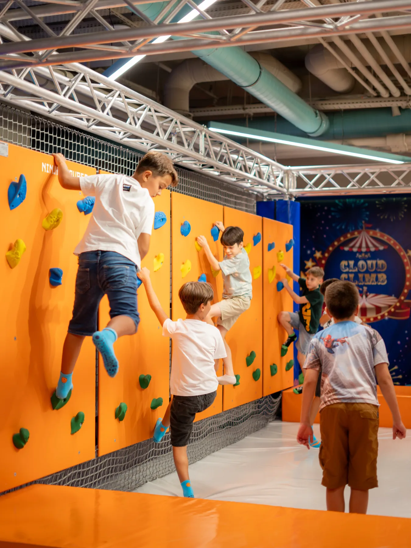 Copii escaladând peretele portocaliu cu prize colorate din zona KLANkids Adventure